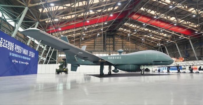 대한항공 부산 테크센터 격납고 내부에 전시된 국산 중고도 정찰용 무인항공기(MUAV) 양산 1호기의 실물 상세 측면 사진. 밝은 조명 아래 웅장한 격납고 천장 트러스 구조를 배경으로, 긴 날개를 펼친 MUAV 1호기가 중앙에 자리 잡고 있다. 회색 동체와 측면에는 ‘중고도정찰용무인항공기(MUAV) 1호기 출고’라는 한글 문구와 함께 로고들이 새겨져 있다. 동체 하단에는 구형 탐지 센서 하우징이 장착되어 있으며, 동체 뒤쪽의 랜딩 기어와 위쪽으로 긴 날개의 구조가 명확하게 보인다. 무인기 왼쪽 뒤로는 다른 군용 차량과 파란색 백드롭이 일부 보인다.