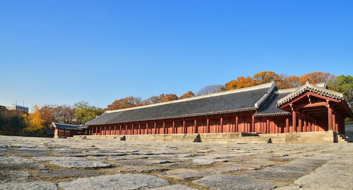 Wide photograph of Jeongjeon Hall at Jongmyo Shrine in Seoul taken from a diagonal angle, showing its long roofline, red wooden columns, and stone courtyard